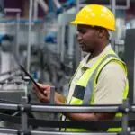 A man wearing a yellow hard hat and reflective vest using a tablet beside curved conveyors and machinery.