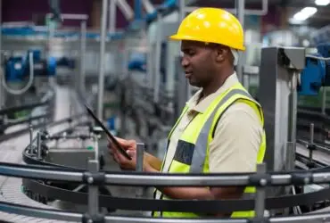 A man wearing a yellow hard hat and reflective vest using a tablet beside curved conveyors and machinery.