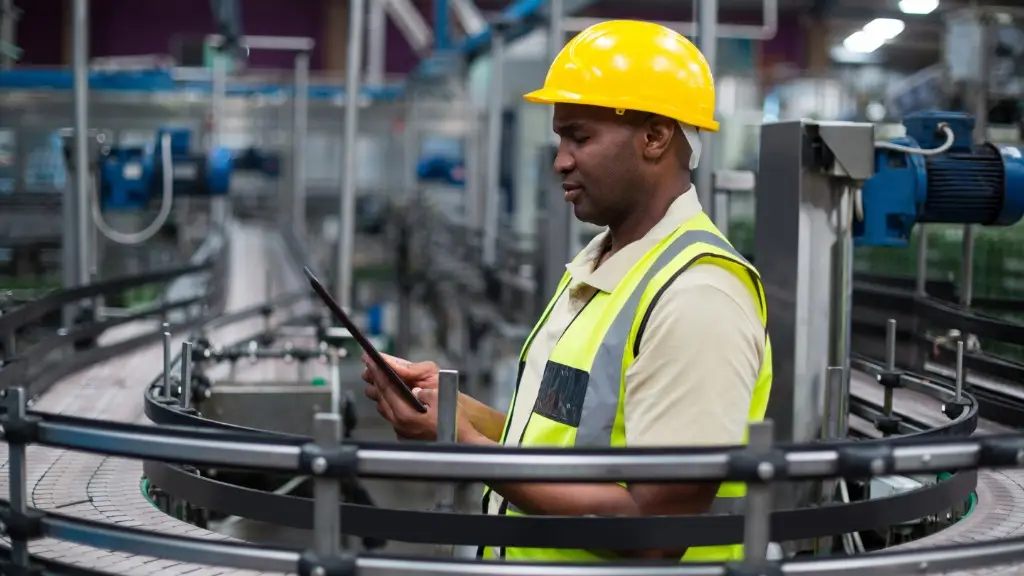 A man wearing a yellow hard hat and reflective vest using a tablet beside curved conveyors and machinery.