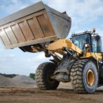 A close-up of an operator driving a large yellow machine with a bucket attachment through an area with sand mounds.