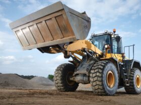A close-up of an operator driving a large yellow machine with a bucket attachment through an area with sand mounds.