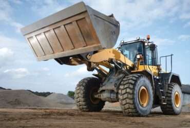 A close-up of an operator driving a large yellow machine with a bucket attachment through an area with sand mounds.