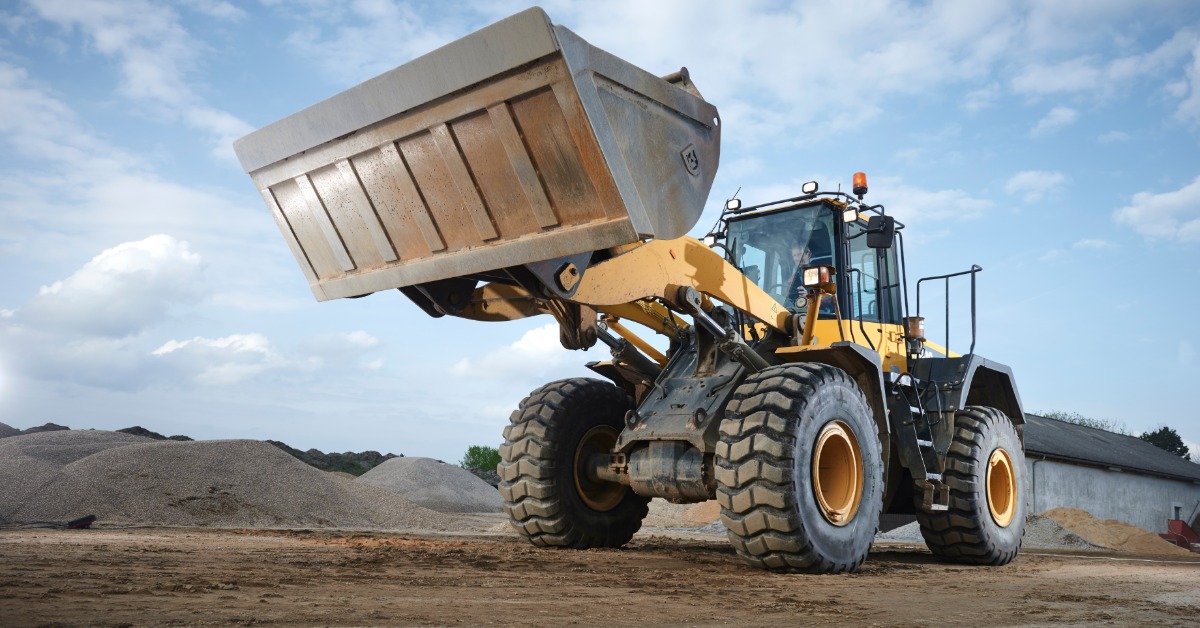 A close-up of an operator driving a large yellow machine with a bucket attachment through an area with sand mounds.