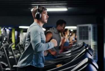 A man wearing headphones running on a treadmill in a gym beside other people using exercise machines.