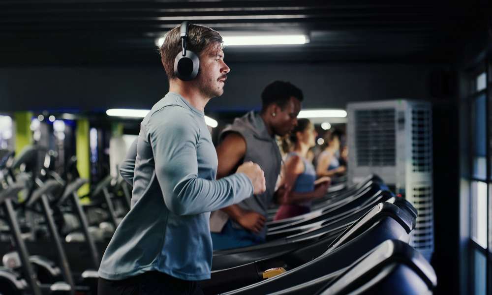 A man wearing headphones running on a treadmill in a gym beside other people using exercise machines.