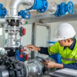 A technician in a hard hat holding a tablet while checking a pressure gauge among industrial pumps, pipes, and valves.