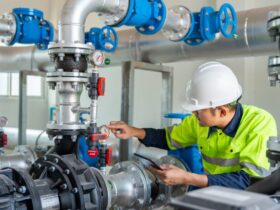 A technician in a hard hat holding a tablet while checking a pressure gauge among industrial pumps, pipes, and valves.