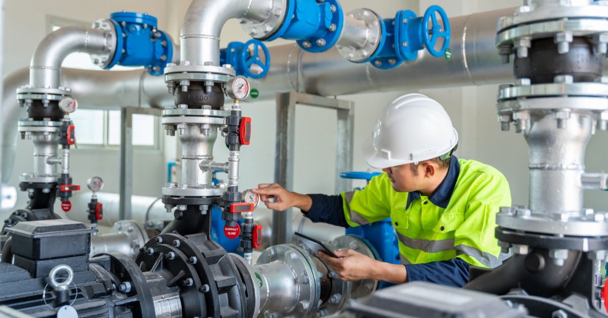 A technician in a hard hat holding a tablet while checking a pressure gauge among industrial pumps, pipes, and valves.