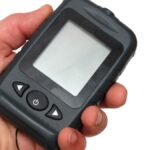 A person is holding a black, portable fishing sonar in front of a white background. The screen is blank.