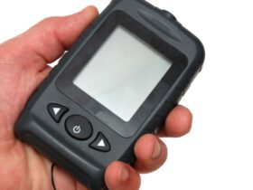 A person is holding a black, portable fishing sonar in front of a white background. The screen is blank.