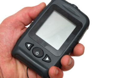 A person is holding a black, portable fishing sonar in front of a white background. The screen is blank.