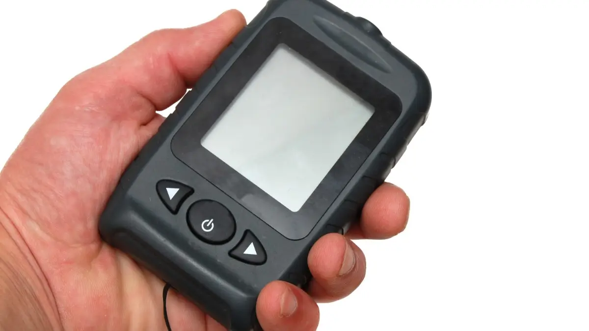 A person is holding a black, portable fishing sonar in front of a white background. The screen is blank.