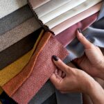 A person compares colorful upholstery fabric swatches in gray, pink, orange, yellow, brown, and neutral tones.