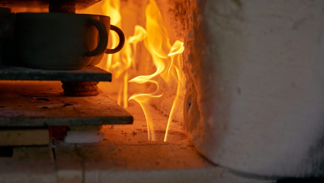 A stack of ceramic cups sit on a flat surface inside a kiln that has a fire going. The flames are orange.