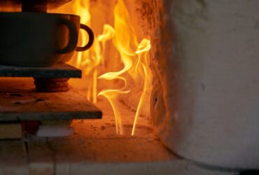 A stack of ceramic cups sit on a flat surface inside a kiln that has a fire going. The flames are orange.