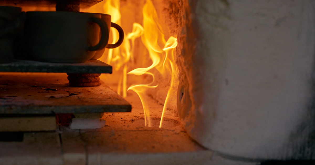 A stack of ceramic cups sit on a flat surface inside a kiln that has a fire going. The flames are orange.