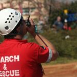 A person is holding a walkie-talkie up to their mouth. The back of the shirt reads "search and rescue."