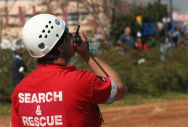 A person is holding a walkie-talkie up to their mouth. The back of the shirt reads "search and rescue."