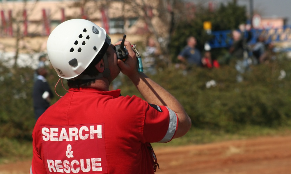 A person is holding a walkie-talkie up to their mouth. The back of the shirt reads "search and rescue."