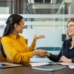 Two smiling women dressed in business attire sit at a conference room table next to each other, shaking hands.