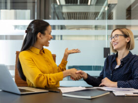 Two smiling women dressed in business attire sit at a conference room table next to each other, shaking hands.