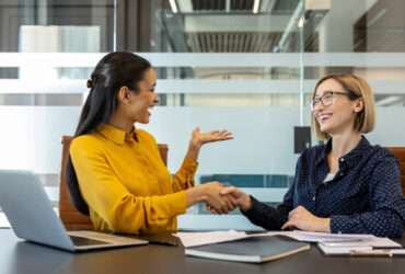 Two smiling women dressed in business attire sit at a conference room table next to each other, shaking hands.