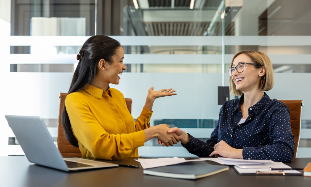 Two smiling women dressed in business attire sit at a conference room table next to each other, shaking hands.
