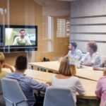 Five men and three women sit around a wooden conference room table, looking at a television screen that displays a person.