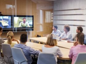 Five men and three women sit around a wooden conference room table, looking at a television screen that displays a person.