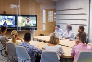 Five men and three women sit around a wooden conference room table, looking at a television screen that displays a person.