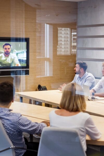 Five men and three women sit around a wooden conference room table, looking at a television screen that displays a person.