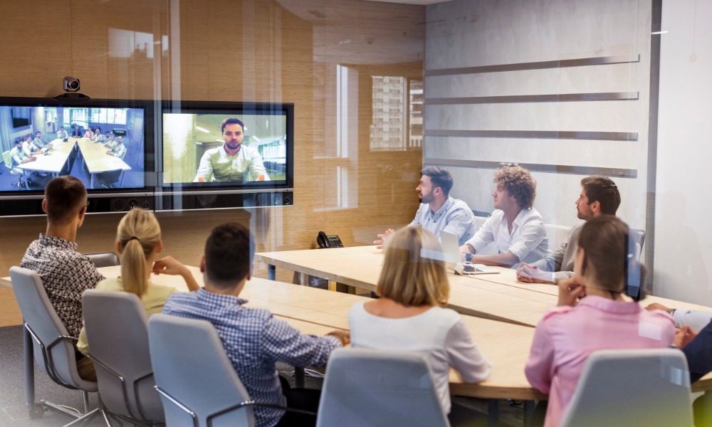 Five men and three women sit around a wooden conference room table, looking at a television screen that displays a person.