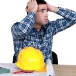 A man sitting at a desk with a hard hat and blueprints in front of him looks in stress at a clock a person is holding.