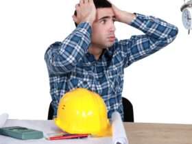 A man sitting at a desk with a hard hat and blueprints in front of him looks in stress at a clock a person is holding.
