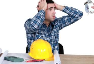 A man sitting at a desk with a hard hat and blueprints in front of him looks in stress at a clock a person is holding.