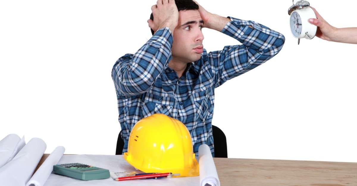 A man sitting at a desk with a hard hat and blueprints in front of him looks in stress at a clock a person is holding.