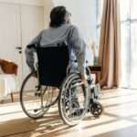 A man in a wheelchair moving towards the large windows within his living room. The space also has a couch.