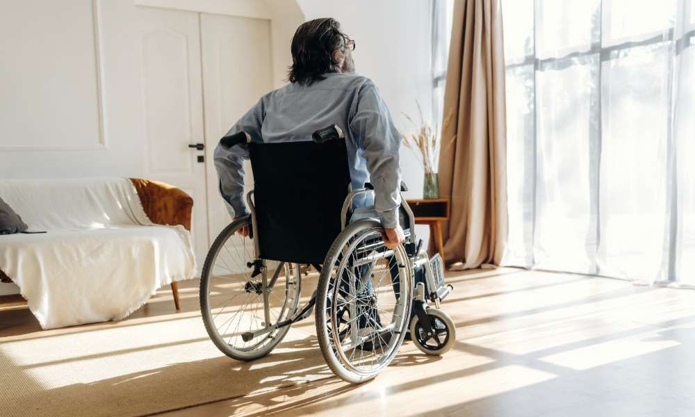 A man in a wheelchair moving towards the large windows within his living room. The space also has a couch.