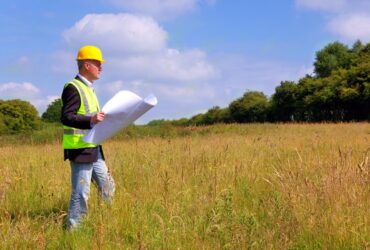 An older man is standing in a field. He’s holding a large piece of paper and wearing a yellow hard hat.