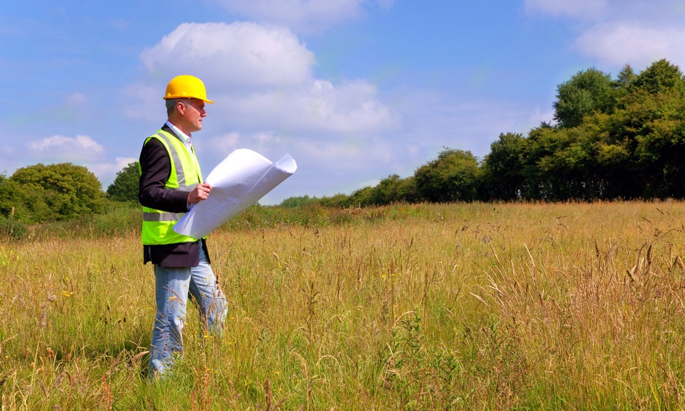 An older man is standing in a field. He’s holding a large piece of paper and wearing a yellow hard hat.