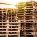 Several tall stacks of wooden pallets sit empty in a warehouse. One stack is on a blue manual forklift.