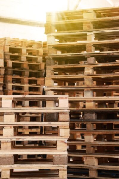 Several tall stacks of wooden pallets sit empty in a warehouse. One stack is on a blue manual forklift.