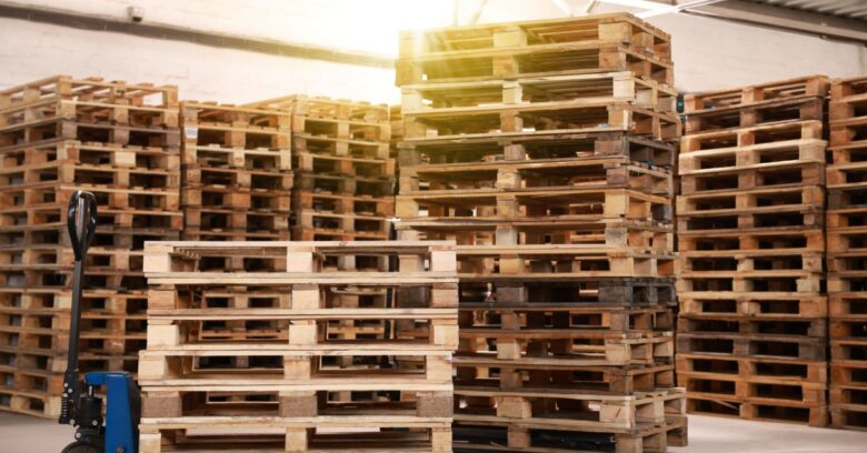 Several tall stacks of wooden pallets sit empty in a warehouse. One stack is on a blue manual forklift.