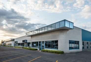 A commercial building has light gray brick walls, blue metal siding, and darkened windows. The parking lot is empty.