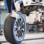 A ground-level view of a man rolling a used car tire in an auto repair shop with a car on a lift in the background.
