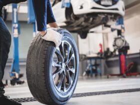A ground-level view of a man rolling a used car tire in an auto repair shop with a car on a lift in the background.