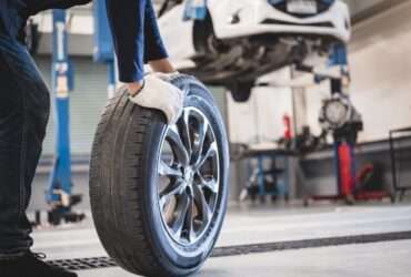 A ground-level view of a man rolling a used car tire in an auto repair shop with a car on a lift in the background.