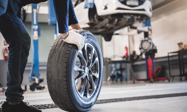 A ground-level view of a man rolling a used car tire in an auto repair shop with a car on a lift in the background.