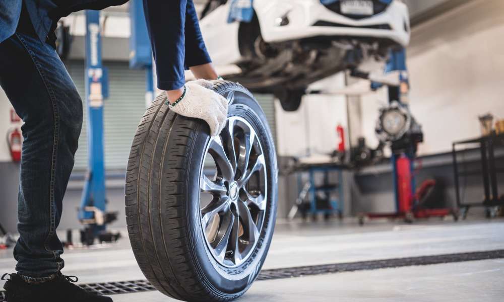 A ground-level view of a man rolling a used car tire in an auto repair shop with a car on a lift in the background.