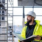 A man in a hard hat is holding a binder and is looking out into the distance while he’s standing in a construction zone.